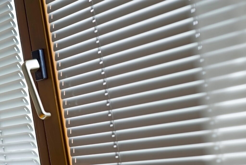 Ein Fenster mit weißem Griff und geschlossenen weißen Lamellen vor braunem Rahmen